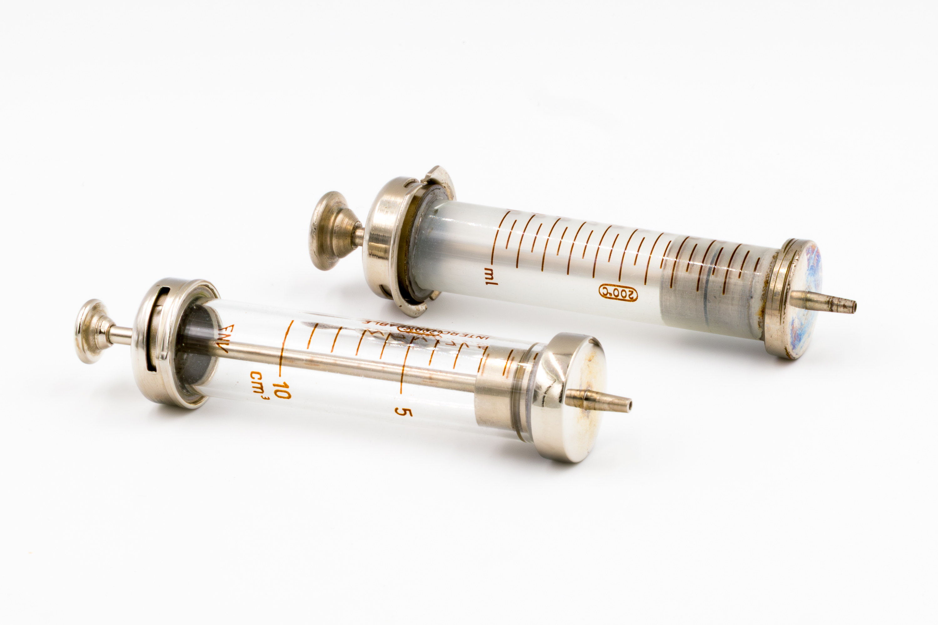 Na zdjęciu znajdują się dwie metalowo-szklane strzykawki typu Luer, wielorazowego użytku, ułożone na białym tle.